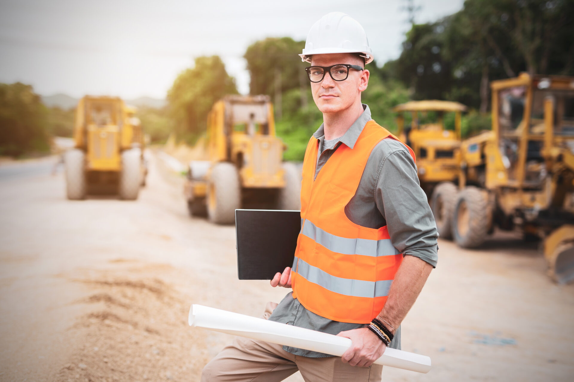 Young engineer holding blueprint and laptop at road construction site.